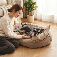 Une femme assise sur un parquet en bois clair, à côté d'un grand lit pour chien de couleur marron où dort un Schnauzer nain. La femme, souriante, tend la main vers le chien pour le caresser. Le lit a des bords surélevés et des détails de boutons. En arrière-plan, un canapé beige, une plante verte et des rideaux.