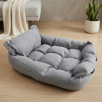 Un lit moelleux pour chien ou chat de couleur grise, doté de bords rembourrés et d'un oreiller intégré, posé sur un parquet en bois clair. L'arrière-plan présente un intérieur lumineux avec un meuble gris sur lequel est drapé un plaid beige, ainsi qu'une plante verte en pot.