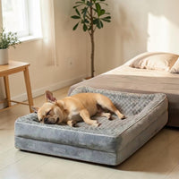 Bouledogue Français fauve profondément endormi dans un panier orthopédique gris bleuté avec surface en peluche douce et rebords rembourrés, posé au pied d'un lit dans une chambre scandinave lumineuse