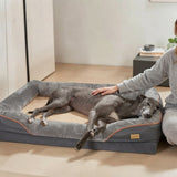 Grand lévrier gris allongé sur un panier orthopédique anthracite avec liseré orange, femme qui le caresse doucement dans un salon épuré et lumineux.
