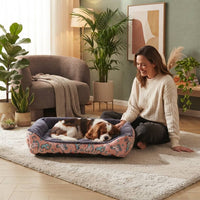 Panier pour chien rose à motifs dans un salon moderne et chaleureux, avec un Cavalier King Charles Spaniel endormi et une femme le caressant.