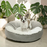Petit chien souriant installé dans un lit orthopédique rond Happy Animalia gris, intérieur en peluche blanche, entouré de plantes vertes.