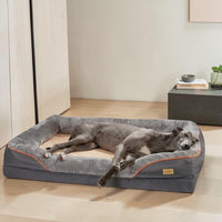 Grand chien allongé et détendu sur un panier orthopédique gris avec rebords rembourrés et liseré orange, posé sur parquet dans un intérieur moderne